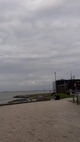 The video shows a waterfront scene on a cloudy day. A group of people is standing by a railing, looking out at the water. There is a brick-paved area in front of them.  A few objects like a trash can and a street lamp can be seen. Some people stroll along the paved area.