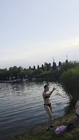 The video opens with a young woman in a black bikini standing on the grassy bank of a body of water. She is facing the water and appears to be interacting with someone off-camera. The water is calm and reflects the sky. In the background, there are people on a dock and some small boats. The scene then shifts to a wider shot of the water and shoreline, showing more reeds and trees. The next shot focuses on a young man standing in the grass, wearing blue swim trunks. He is looking down at his hands, which are holding something. The final shot shows a group of people in the distance at what appears to be a beach or swimming area. The overall environment is a natural outdoor setting, likely a lake or pond, with a clear sky.