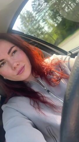 A woman with red hair is driving a car. She is wearing a light gray jacket and has her hand on the steering wheel. The sun is shining through the window. She is looking up and slightly to the right. Trees can be seen through the car window.
