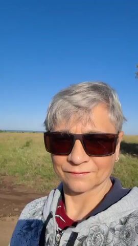 A woman with short gray hair and sunglasses is taking a selfie outdoors on a sunny day. She is wearing a gray hoodie with a floral pattern. The background shows a dirt path, grassy fields, and a clear blue sky. There is a tree on the left side of the frame.