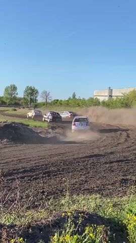 This dynamic image captures a multi-car rallycross or dirt track race in progress, set against a bright, clear daytime sky. Several cars are depicted in motion, kicking up significant clouds of dust as they navigate a wide, unpaved track. The vehicles appear to be modified hatchbacks, commonly used in such events.

In the foreground, a white and blue car with the word "POLO" clearly visible on its rear leads the pack, its wheels turning and exhaust visible. Behind it, at least three other cars are closely following, also raising plumes of dirt. One of these trailing cars is dark-colored, and another is lighter-colored. The cars are rounding a left-hand bend in the track, emphasizing the high-speed action of the event.

The setting is an outdoor, purpose-built dirt racing circuit. The track itself features elevated banks and turns, indicative of a challenging course. In the background, green trees line the horizon, and a large, light-colored industrial-style building is visible, suggesting the race takes place in a semi-rural or industrial area. The weather is sunny and dry, contributing to the substantial dust clouds. The scene is located in Kanjiza Municipality, Serbia. No specific individuals are clearly identifiable, but the focus is on the competitive and energetic nature of the car race.