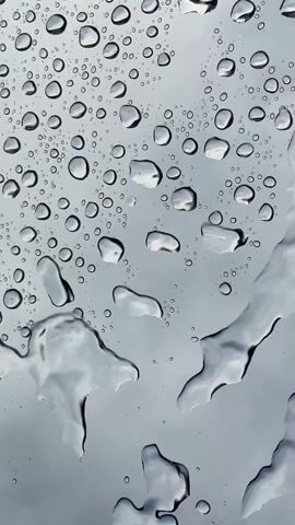 The video shows a close-up view of raindrops on a glass surface. The raindrops vary in size, some are large and elongated while others are small and circular. The background is a grey, overcast sky, partially visible through the water droplets. The camera angle appears to be looking upwards through the glass.  The raindrops slowly slide down the surface as the video progresses.