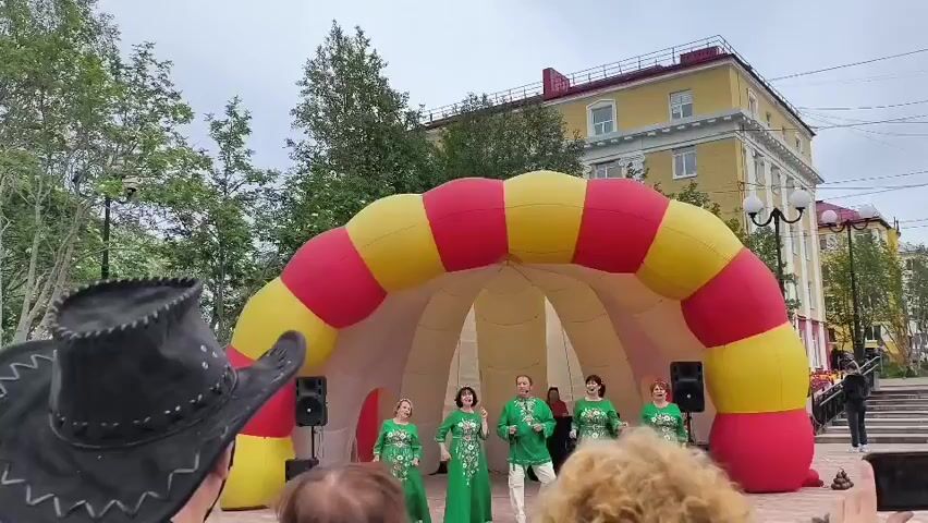A group of five people, four women and one man, dressed in matching green traditional outfits with ornate white embroidery, are performing on a temporary outdoor stage. They are positioned in front of a large, inflatable, dome-shaped structure with alternating red and yellow striped segments. The stage appears to be in an open public area, possibly a square or park, with trees visible in the background. To the left, the back of a person wearing a black cowboy hat is visible, facing the performance. To the right, a yellow building with multiple windows stands behind the inflatable structure, and a lamppost is visible near it. Two black speakers are positioned on either side of the inflatable entrance. The sky is overcast, suggesting a cloudy day. The people on stage are actively engaged in what appears to be a song and dance performance.