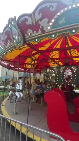 This media file shows a carousel with several horses. The carousel has a red and yellow striped canopy. The horses are various colors and are attached to poles that allow them to move up and down. The scene appears to be outdoors, possibly at a fair or amusement park. There are some buildings and fencing visible in the background. The weather appears to be overcast, and the time of day is likely daytime. There is a railing in the foreground of the image. The carousel itself is ornate, with decorative trim and lights. The visible text on the carousel appears to read "FS" with some decorative elements. The overall impression is that of a classic carnival ride.