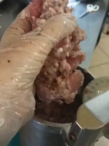 A gloved hand is shown preparing ground meat inside a metal container. The hand is pressing down on the meat, which appears to be seasoned with small dark specks. In the background, other metal bowls and a pair of scissors can be seen on a stainless steel surface.