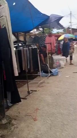 The video shows an outdoor market stall with various clothing items displayed on racks. The stall is covered by a blue tarp and an umbrella. People are seen browsing the merchandise and interacting with each other. Some of the clothing items appear to be used or second-hand. The ground is paved with concrete. The scene suggests a busy market day with potential customers examining the goods for sale.