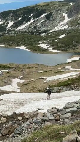 A person wearing a white hat, a dark jacket, and shorts walks down a snowy slope. They are carrying a backpack. The scene is a mountainous landscape with a lake in the background. The mountains are green and covered in patches of snow. The lake is blue and calm. The foreground is rocky with some grass. The sky is partly cloudy. The time of day appears to be daytime, and the weather is likely cool due to the snow. The location is Panichishte, Bulgaria. There is no visible text in the media file.