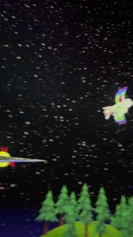 The video displays a projection of a starry night sky with animated creatures flying across it. In the foreground, a stylized landscape of green trees and colorful mountains is visible. Several people are present, with their silhouettes or partially visible figures in the lower portion of the frame, suggesting they are viewing the projection. The animated objects include a white bird with a rainbow tail, a rocket-shaped object with red and blue fins, a yellow bird, and several colorful fish-like creatures. The projection appears to be on a large curved surface, possibly a wall or dome. The overall atmosphere is whimsical and fantastical.