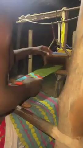 A man is weaving fabric on a traditional loom. He is shirtless and the loom appears to be made of wood. He uses a shuttle to pass yarn through the warp threads, creating a colorful pattern. The woven fabric is visible beneath the working area. The environment seems rustic, with wooden beams and simple lighting.