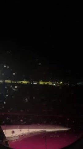 The video, filmed at night from an airplane window, shows a wing with "NO STEP" markings visible on its surface. In the distance, an airport or industrial area with lights is illuminated against the dark sky. The window is speckled with water droplets or condensation, distorting the view slightly.