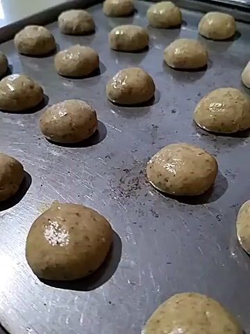 A close-up, high-angle shot shows a baking sheet filled with uncooked, round, light brown cookie dough balls. The dough balls are evenly spaced and appear to have small dark flecks within them, possibly herbs or spices. Some of the dough balls are slightly shiny, suggesting they may have been brushed with an egg wash or oil. The baking sheet is a metallic, grey color with some scratches and marks, indicating it is well-used. The background is blurred, but appears to be a kitchen counter or workspace. The lighting is bright and direct, casting subtle shadows on the dough balls and the baking sheet. The video is static, with no apparent camera movement or subject motion.