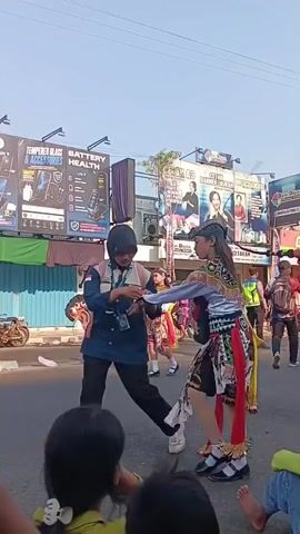 The video shows a street parade with people in traditional costumes, some riding on what appear to be horse-like puppets. In the foreground, two children are visible from behind, watching the parade. On the left, a person in a blue jacket and black pants with a name tag is adjusting something on their wrist. The main focus is on a person in a purple top and a patterned skirt, holding a large, black, horse-like puppet with red and white decorations. This person is walking and turning, with the puppet appearing to move with them. Other participants in similar traditional attire, some with red sashes, are also visible further down the street, moving in the same direction. In the background, there are buildings with signs, including one for "LCD," "TEMPERED GLASS & ACCESSORIES," and "BATTERY HEALTH." There are also people watching from the sides of the street and some motorcycles parked. The sky is bright and clear, suggesting it is daytime. The overall atmosphere appears to be festive, with a cultural performance taking place.