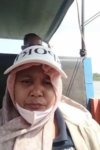 The video shows two people on a boat. The person in the foreground is a woman wearing a headscarf, a face mask, and a baseball cap that says "SPORT". She is looking at the camera. Behind her, a man wearing sunglasses is visible. The boat appears to be on a river or body of water, with greenery and trees visible on the banks. The weather seems to be cloudy.