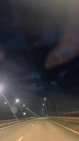 The video shows a view from a vehicle traveling on a highway at night. Streetlights illuminate the road and the sky above is dark with some clouds. The road appears to be a multi-lane highway with yellow and white lane markings. There are no other vehicles visible in the immediate vicinity, and no people are visible. The scene conveys a sense of nighttime travel on an open road.