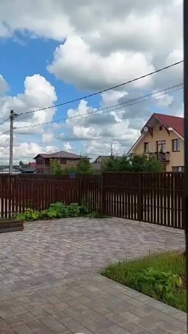A brief, upward-facing video captures a residential outdoor scene on a cloudy day. The foreground is dominated by a paved courtyard, laid with rectangular interlocking stones in varying shades of gray and tan. To the left, a dark wooden planter box rests on the edge of the courtyard. A dark brown wooden fence, with vertical slats and a horizontal top rail, encloses the courtyard and extends across the frame. Beyond the fence, several houses with red roofs are visible, interspersed with green trees and foliage. A utility pole with power lines stretches across the sky. The sky itself is a vibrant blue, punctuated by large, fluffy white cumulus clouds. The camera angle is static, looking up from a low vantage point.