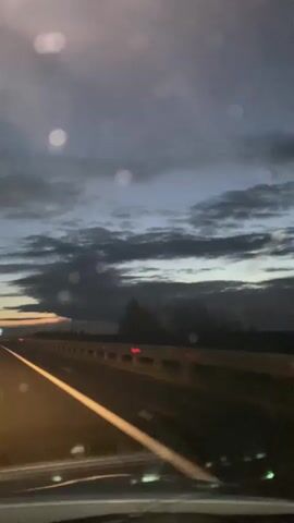 The video shows a view from a vehicle traveling on a highway at dusk or dawn. The sky is filled with dark clouds, with a hint of orange and yellow light on the horizon. The road ahead is visible, with a white line marking the lane. There are some lights in the distance, possibly from other vehicles or buildings. The video is shaky, suggesting it was recorded while the vehicle was in motion. There are also some circular blurs in the video, which could be water droplets on the windshield or lens flares.