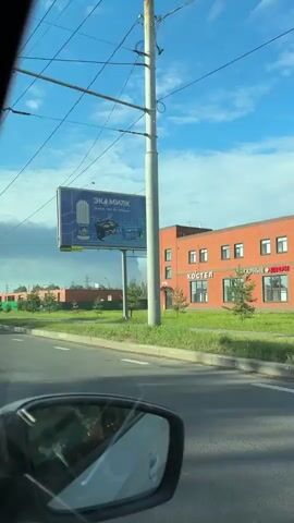 The video shows a view from inside a car looking forward and slightly to the right. The car is on a road with a lane marker and a white dashed line. On the right side of the road, there is a sidewalk, a green fence, and several buildings under construction. One building is a brick building with orange bricks and windows. Another building appears to be a concrete structure with exposed rebar. A large billboard with Russian text and images is visible on a pole to the right. The billboard features an advertisement for "ЭКАМИЛК" (Ekamilk). Further in the background, there is a large, dark cloud formation in the sky, possibly smoke from a fire. Overhead, there are power lines and utility poles. A street sign with directions to "КОР. РОГА" and "МОСКВА" is visible above the road. The sky is blue with some scattered white clouds. The car's side mirror is visible in the lower left corner of the frame. The video appears to be shot during the daytime.