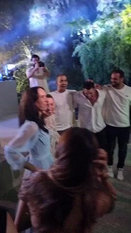 The video shows an outdoor evening event with a group of people dancing. In the foreground, several women are visible from behind, with one woman in a light blue dress on the left. In the center, a couple is dancing, with the woman wearing a white lace dress and the man in a white button-down shirt and black pants. To their right, two men in white shirts and dark pants are also dancing and interacting. In the background, a man with a beard is standing on a platform, holding a camera and appearing to film the event. Stage lighting with purple and pink hues illuminates the scene, creating a smoky atmosphere, and trees are visible in the background. Some of the people are smiling and appear to be enjoying themselves. The ground is paved, possibly with gravel or stone.