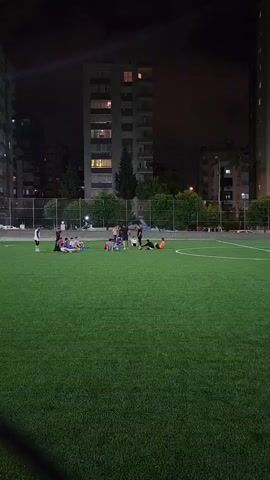 A group of young people are gathered on a brightly lit artificial turf soccer field at night, with apartment buildings visible in the background. Some individuals are sitting on the grass in a huddle, while others are standing or lying down. The overall atmosphere appears casual and relaxed, possibly indicating a break or the end of an activity.