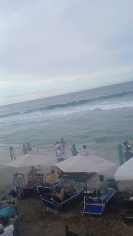 The video shows a crowded beach. Many people are sitting under umbrellas and on beach chairs. Others are walking along the shoreline or standing in the water. Waves are visible in the ocean. There is a tall white flag with writing on it standing in the sand near the water's edge. The sky is overcast.