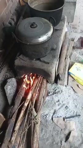 A video shows a traditional wood-burning stove with a pot on top. The fire is burning brightly, with flames visible through the opening of the stove. The stove is made of concrete or clay, and there are several pieces of firewood stacked beneath it, some of which are burning. The pot on top appears to be made of metal and has a lid. The background shows a brick wall. The ground around the stove is covered in ash and debris.