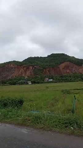 This media file appears to be a collection of short video clips shot from a moving vehicle, likely a car, on a cloudy day in Lampisang, Indonesia. The primary subject is the scenery of the Indonesian countryside, characterized by lush green rice fields, rolling hills, and a large, reddish-brown cliff face in the background.

Several small, makeshift roadside stalls selling drinks and snacks are visible. One stall prominently features an advertisement for "Jambu Sari Dek Lia," which translates to "Sari Guava Dek Lia," implying it sells guava juice or related products. Another stall has text that appears to read "TEH GORENG," potentially indicating "fried tea" or a special type of tea preparation. Small huts, likely used by farmers for shelter in the fields, are also scattered across the landscape.

The weather appears overcast and possibly humid, typical of tropical regions. The time of day is not precisely discernible but seems to be daytime. The overall mood conveyed is one of travel through a scenic, rural area. The clips capture the natural beauty of the landscape with interspersed glimpses of local commerce along the roadside. The movement of the vehicle suggests a journey or a leisurely drive through the area.