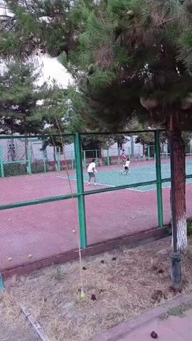 The video shows a tennis court with three people. Two men are playing tennis on the court, while a third person, a young boy, is also on the court. One man is wearing a white shirt and shorts, and is holding a tennis racket. The other man is wearing a red shirt and shorts, and is also holding a tennis racket. The young boy is wearing a white shirt and shorts. There are tennis balls scattered around the court. The court is surrounded by a fence. In the background, there is a building and trees.