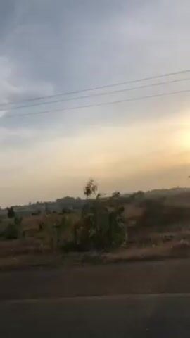 The video shows a sunrise viewed from a moving vehicle on a road. The sun is low on the horizon and obscured by trees and utility poles. The landscape includes dry grass and scattered trees. The sky is a mix of blue and orange hues.