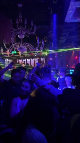 The video shows a crowded dance floor at a nightclub. People are dancing under flashing blue and red lights, and green laser beams move across the room. There's a large white chandelier hanging from the ceiling. The crowd is a mix of men and women, mostly wearing dark clothing. Some people have their arms raised as they dance. The overall atmosphere is energetic and festive.
