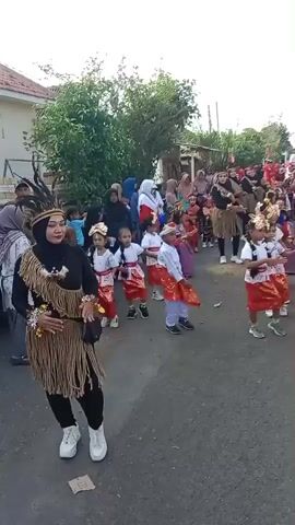 A group of children and adults are walking down a street. The children are dressed in traditional outfits, and some of them are dancing. The adults are watching the children and some are also participating in the procession. The street is lined with houses and trees. The sky is clear and the sun is shining. The video appears to be taken from a moving vehicle. The people are enjoying themselves and there is a festive atmosphere.
