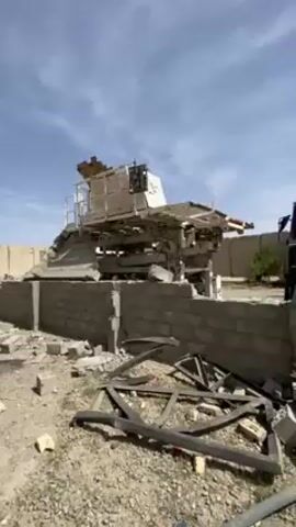 The video shows a scene of devastation and destruction with debris and rubble scattered across the ground. A large white machine or structure sits atop a partially destroyed cinder block wall. A damaged truck is visible behind the wall, with a shipping container nearby. As the camera pans out, several men dressed in traditional clothing are seen walking through the rubble. The sky is overcast with patches of blue. The overall impression is one of a recent destructive event.