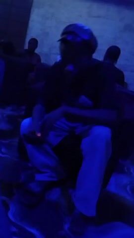 A video taken indoors under blue light shows a person sitting in a chair and appearing to manipulate something in their hands. The individual is wearing a dark t-shirt and light-colored pants. There are other people in the background, further back in the room. The floor seems to have a pattern or design under the blue light.