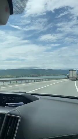 A car drives along a highway next to a wide body of water, with mountains visible in the distance under a cloudy sky. The dashboard of the car is visible in the foreground, showing a digital display with "15:34," "19.0," "22.0," and "21C," along with "Apple CarPlay." A blue Volvo truck carrying multiple cars passes in the opposite direction, followed by a white Dacia car. A metal guardrail separates the road from the water. The scene suggests a daytime drive on a scenic route, likely alongside the Danube River near Drobeta-Turnu Severin, Romania. Voices can be heard from inside the car.