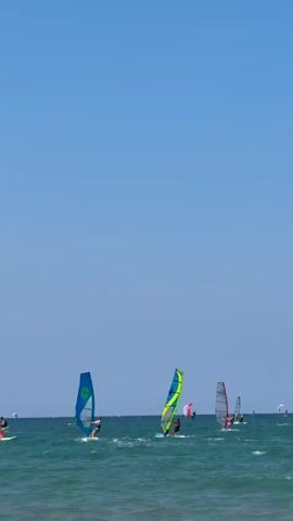 The video shows a clear blue sky above a turquoise ocean with moderate waves. Several people are windsurfing on the water. In the foreground and mid-ground, there are at least five windsurfers, each with a colorful sail, actively maneuvering their boards. The windsurfers are spread out across the frame, with some closer to the camera than others. The overall impression is one of a sunny day with enjoyable windsurfing conditions.