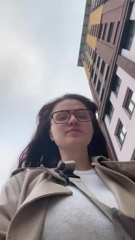 A woman in a beige trench coat, a white t-shirt, light blue jeans, and white sneakers walks along a beige brick sidewalk. She wears brown-framed glasses and has brown wavy hair. She is wearing a black strap for a bag across her chest. In the beginning, the camera is aimed up at a tall, multi-colored building. The camera rotates and is now aimed at her face as she walks forward. There is background noise and Russian spoken in the background.