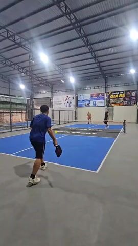 Two men and two women are playing pickleball indoors on a blue court with a gray floor. The court is divided by a net. The players are wearing athletic clothing. One man in a blue shirt is in the foreground, facing away from the camera, and appears to be hitting the ball. Two other players are on the opposite side of the net, and another player is on the same side as the man in the blue shirt. The court is lit by overhead lights. There are advertisements on the wall behind the court.