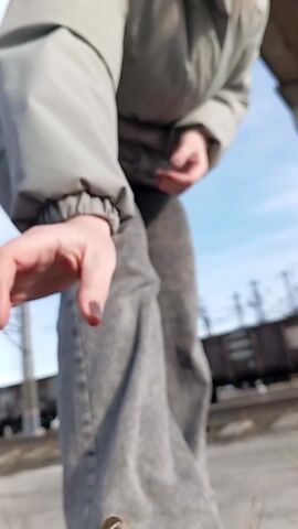 A woman with red hair, dressed in a grey jacket and grey jeans, is standing outdoors. The background includes railway tracks, a freight train, power lines, and a blue sky with some clouds. The woman is adjusting her hair, pushing it back from her face. The camera appears to be positioned low and looking upwards.