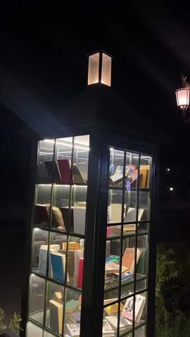 The video shows a tall, narrow, glass-enclosed structure resembling a telephone booth, filled with books and illuminated by internal lights. The structure is dark green and has a lantern-like light fixture on top. The books are of various sizes, colors, and bindings, and are arranged on shelves within the glass panes. Some books are standing upright, while others are lying down. The background is dark, suggesting it is nighttime. There are no people visible in the video, and no actions or interactions are taking place other than the subtle flickering of the lights, which may be due to the camera's refresh rate or a slight fluctuation in the lighting. The environment appears to be outdoors, with some greenery visible on the right side of the frame.
