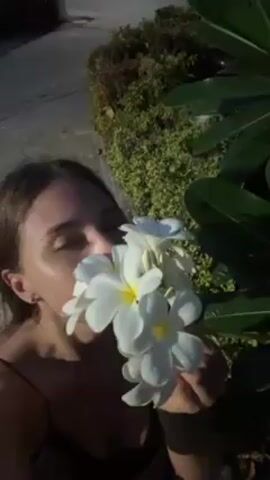 A woman with long brown hair is standing outside and smelling white flowers with yellow centers. She is wearing a black top. Behind her is a bush and a sidewalk. Sunlight is shining on the scene. The woman closes her eyes and takes a deep breath as she smells the flowers.