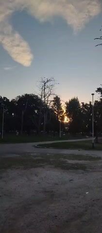 The video pans upwards from a gravel patch in a park toward a cloudy sky at dusk. The sun is setting behind a line of trees, creating a silhouette effect. The park has a paved path with a small grassy area in the middle. A person in a yellow shirt is visible in the distance. There are several bare trees mixed in with the foliage. The sky is a pale blue with a long, thin cloud. Streetlights are spaced along the path.