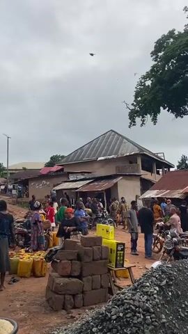 The video shows a bustling outdoor scene in a rural area, likely a village, filled with people and activity. The ground is reddish-brown dirt and gravel, with a mix of parked motorcycles and larger vehicles like vans and a small truck scattered throughout. Many individuals are carrying or surrounded by yellow plastic jerrycans, suggesting they are collecting or distributing some type of liquid, possibly water or fuel. A water tower stands prominently in the middle of the scene, adding to the impression of water scarcity or a centralized water source. The buildings in the background are simple structures with corrugated metal roofs. The sky is overcast, and the surrounding area has lush green trees. The general atmosphere is one of communal activity and resourcefulness, with a sense of people going about their daily routines.