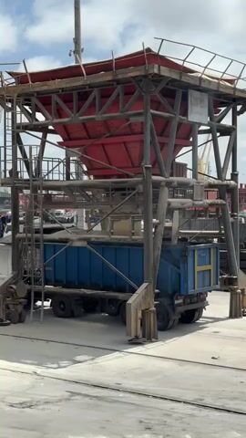 The media file captures a loading operation at a port in Casablanca, Morocco. The primary subjects are industrial equipment and a truck. A large, red, funnel-shaped hopper, supported by a red and grey metal structure, dominates the upper portion of the frame. Below the hopper, a blue truck with a large cargo bed is positioned to receive material. The truck has multiple axles and appears to be a heavy-duty vehicle.

The activity taking place is the transfer of bulk material, likely from the hopper into the truck's cargo bed. The hopper is full, suggesting material is being dispensed or is about to be dispensed. The scene is set in an outdoor industrial area, typical of a port, with concrete ground and other industrial structures or equipment visible in the background, including cranes and what appear to be shipping containers. The sky is cloudy, indicating a day with mixed weather conditions, not necessarily bright sunshine.

There are no people clearly visible in the foreground or interacting directly with the loading process. The time of day appears to be daytime, given the lighting. A visible sign on the structure reads "MARSA MOROCCO" and below it "000006". The overall impression is one of industrial activity and logistics, focused on the efficient movement of goods. There are no overt emotional cues conveyed by the scene, as it is primarily focused on mechanical processes.