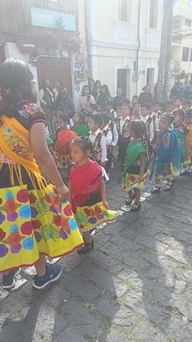A group of young children are walking in a parade on a cobblestone street. The children are wearing traditional clothing and holding hands. A woman in a colorful dress is leading the children. There are buildings and spectators on both sides of the street.