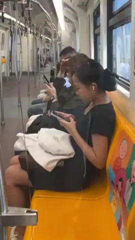 The camera shows the inside of a subway car with a row of passengers sitting on an orange bench seat. Each passenger is engrossed in their cell phone. The lighting is bright and diffused. Some of the subway windows are visible, showing the outside. Some handrails hang from the ceiling of the subway car.
