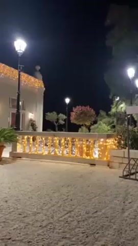 At night, the video shows a building decorated with lights.  The building appears to be white, with a string of yellow lights hanging from its roof.  There are windows visible, some with white frames. In front of the building is a stone railing also adorned with yellow lights.  The ground is a light-colored stone or gravel. There are potted plants nearby. The scene is illuminated by street lights. The camera appears to be inside a vehicle, moving slowly towards the building.  The focus stays on the building and its decorative lighting.