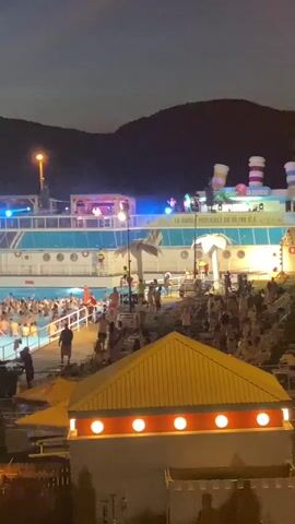 The video shows a nighttime scene at a crowded outdoor pool party. The pool is filled with many people, mostly young adults, some of whom are swimming or standing in the water. The pool area is decorated with lights and palm trees. In the background, there is a large structure resembling a ship, also illuminated with lights, with a stage or platform on top where people appear to be performing or DJing. The overall atmosphere suggests a lively event, possibly a concert or a festival, taking place after dark. Hills or mountains are visible in the far background against the night sky. Several smaller structures, possibly food stalls or seating areas, are also present in the foreground, illuminated with yellow and orange lights. The lighting is primarily artificial, with a mix of colorful party lights and functional illumination.
