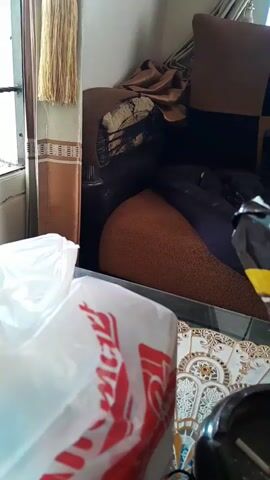 The video shows a close-up of a table with several items on it. In the foreground, a white plastic bag with red lettering is visible. To the right of the bag is a black ashtray containing a lit cigarette, ash, and cigarette butts. Behind the ashtray is a partially opened bag of chips with yellow and black packaging. In the background, a portion of a couch with a checkered pattern of brown and black squares can be seen, along with a black plastic bag draped over it. The lighting suggests an indoor environment. The video is shaky and appears to be handheld.