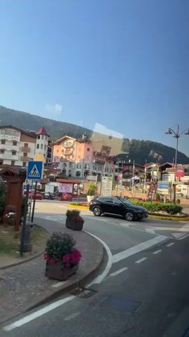 A black SUV drives around a roundabout in a town. Buildings with colorful facades are visible on the left side of the road. A man is walking on the sidewalk. There is a street lamp and various flags on poles. The sky is clear and blue.
