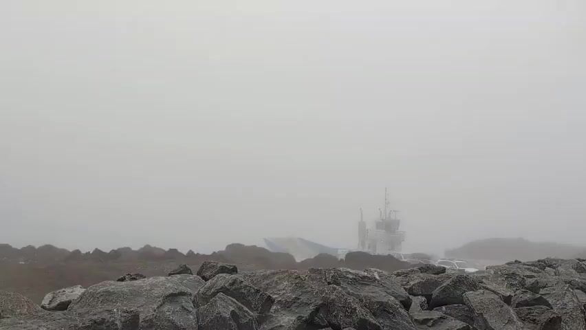 The video shows a wide shot of a foggy environment. In the foreground, there are large rocks. In the middle ground, a ferryboat is visible through the fog. The ferry has a white cabin structure with a radar mast on top. The deck of the ferry appears to be flat and blue and yellow markings can be seen. There are vehicles on the ferry, though they are indistinct due to the fog. The background is a solid wall of white fog. The overall scene conveys a sense of limited visibility and a quiet, perhaps isolated, atmosphere.
