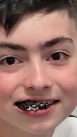 A close up shot shows a young man with brown hair and brown eyes smiling with crushed Oreo cookies between his teeth. The background is blurry.